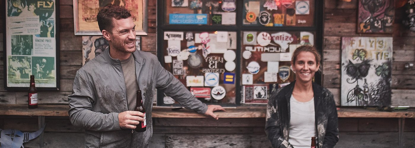 Two people smiling and holding beer bottles in front of a wall covered with posters and stickers.