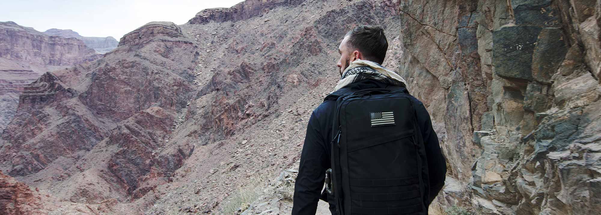 Man with a black backpack and scarf hiking in a rocky canyon landscape.