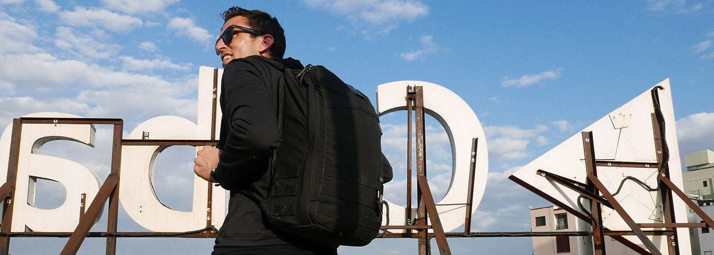 Person in sunglasses and black jacket with backpack, standing on a rooftop near a large, partially obscured sign.