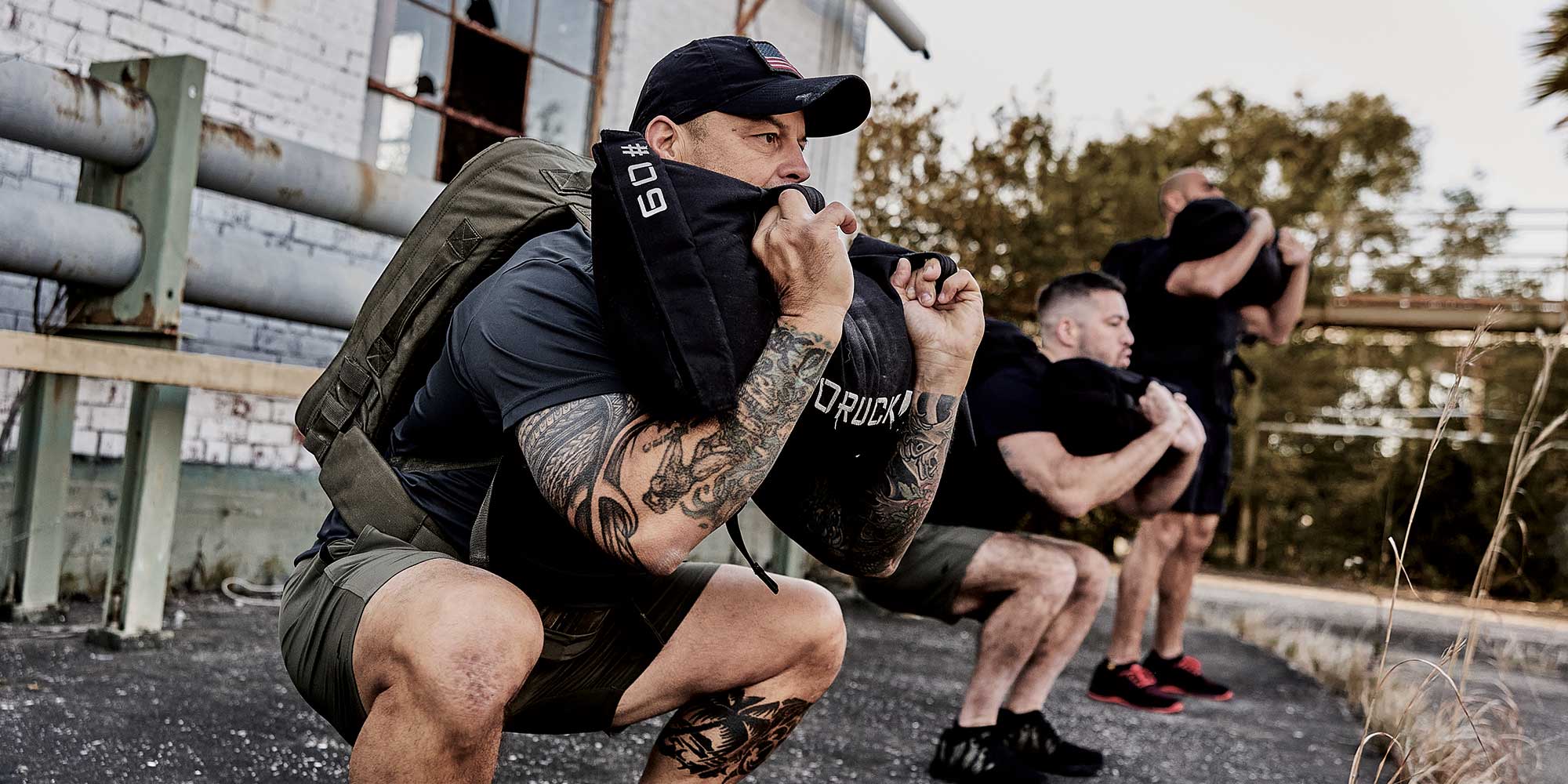 Three men squatting outdoors with weighted bags, showing intense concentration and strength.