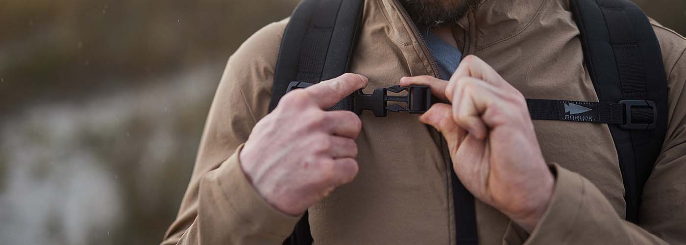 Person fastening a chest strap on a backpack while wearing a tan jacket outdoors.