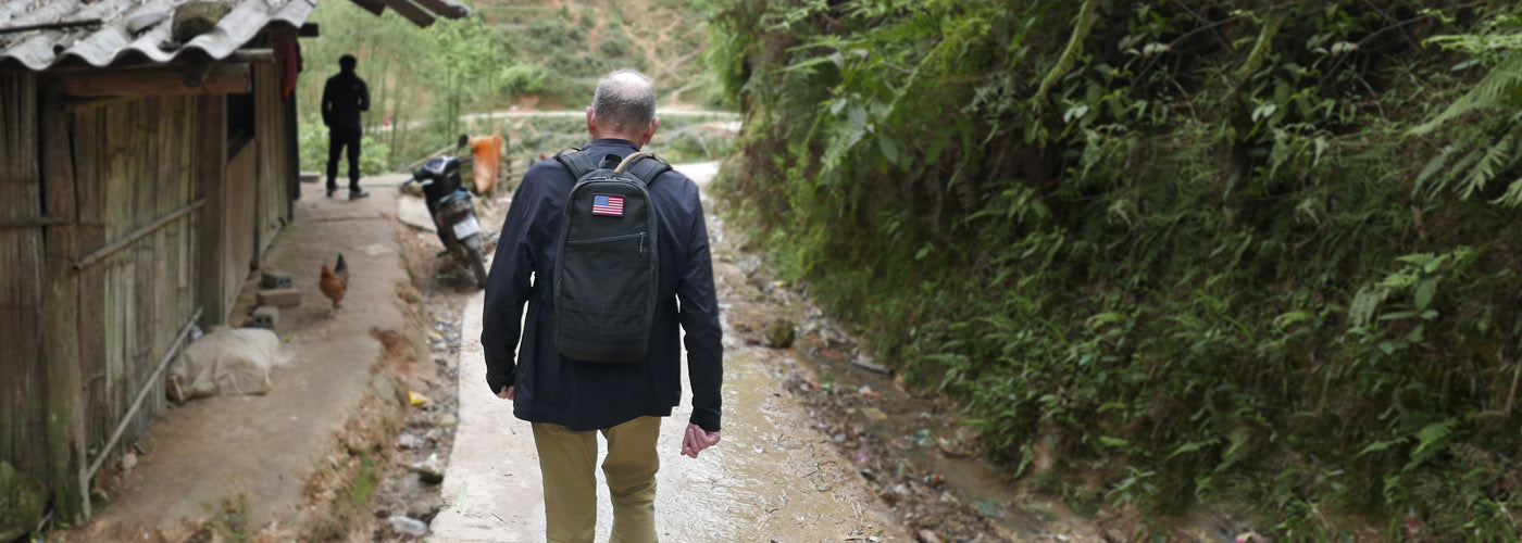 A person with a backpack walking on a dirt path beside a small stream and a house, with greenery on both sides.