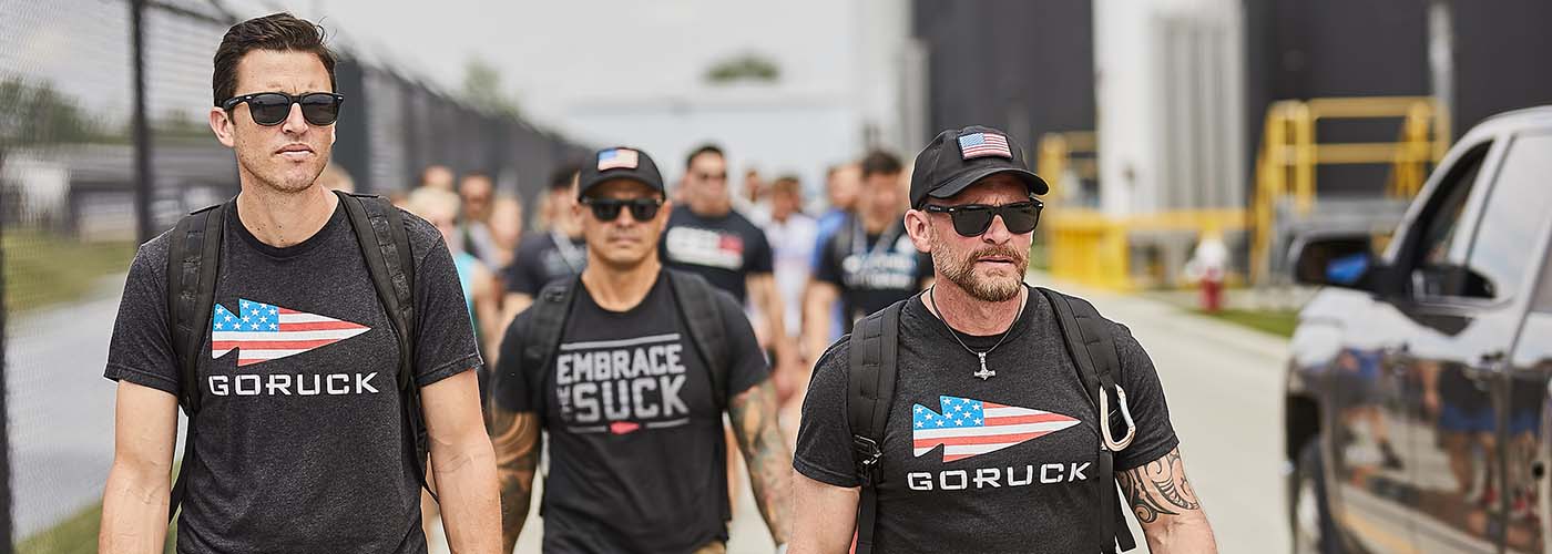 Three men in black GORUCK shirts and sunglasses walk outside, carrying backpacks.