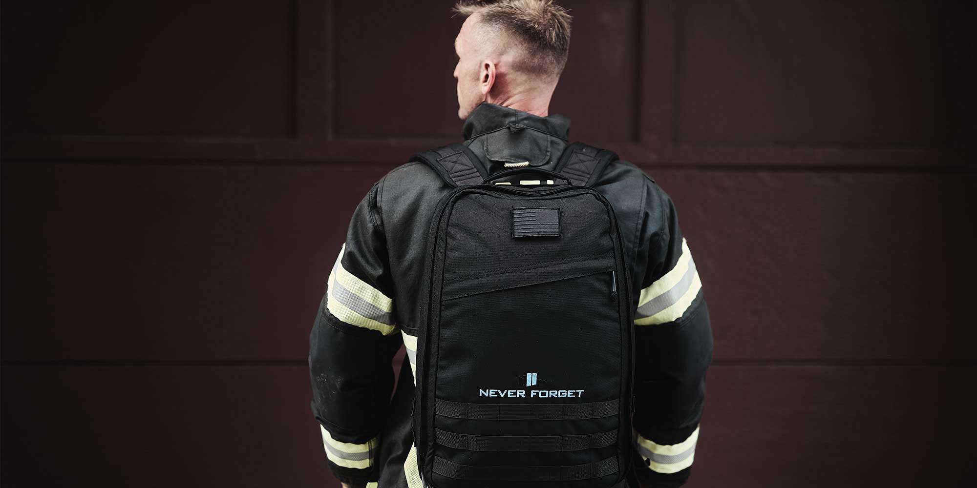 A firefighter in uniform faces away, wearing a black backpack with "Never Forget" written on it.