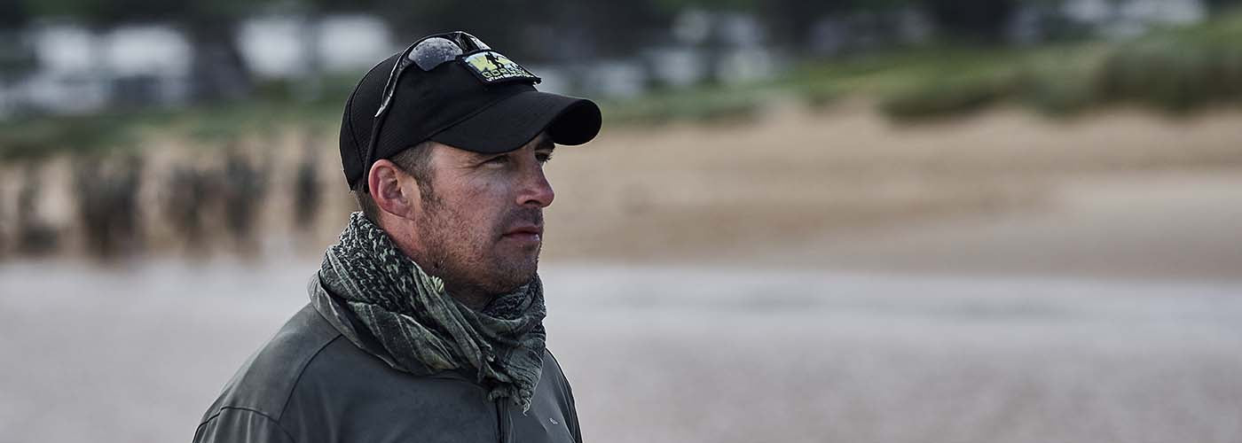 Man in cap and scarf stands on a beach, looking intently. Blurred figures in background suggest a group or team activity.