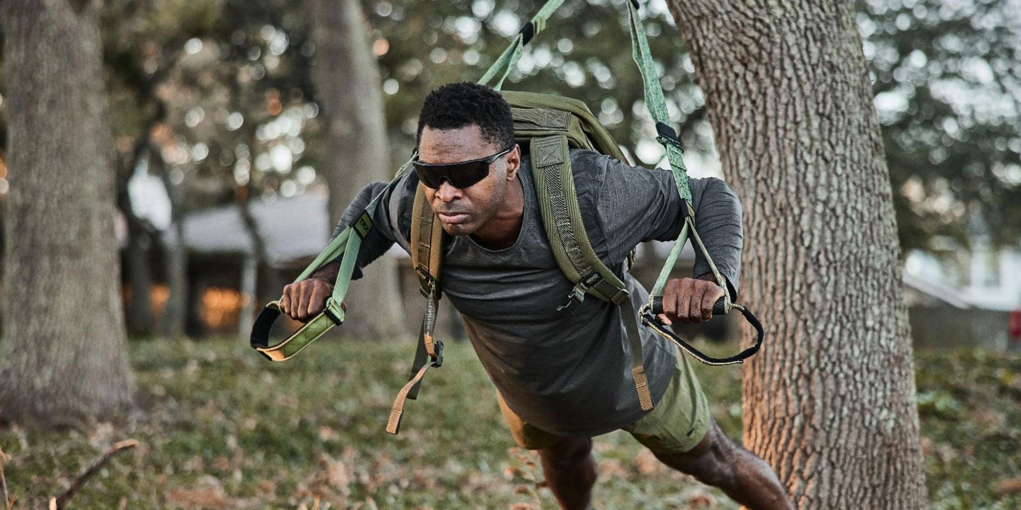 A man exercises outdoors with suspension trainers, wearing sunglasses and a backpack.