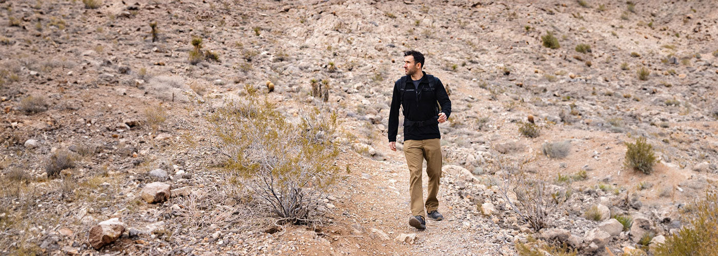 Person walking on a rocky desert path, surrounded by sparse shrubs and dry landscape.