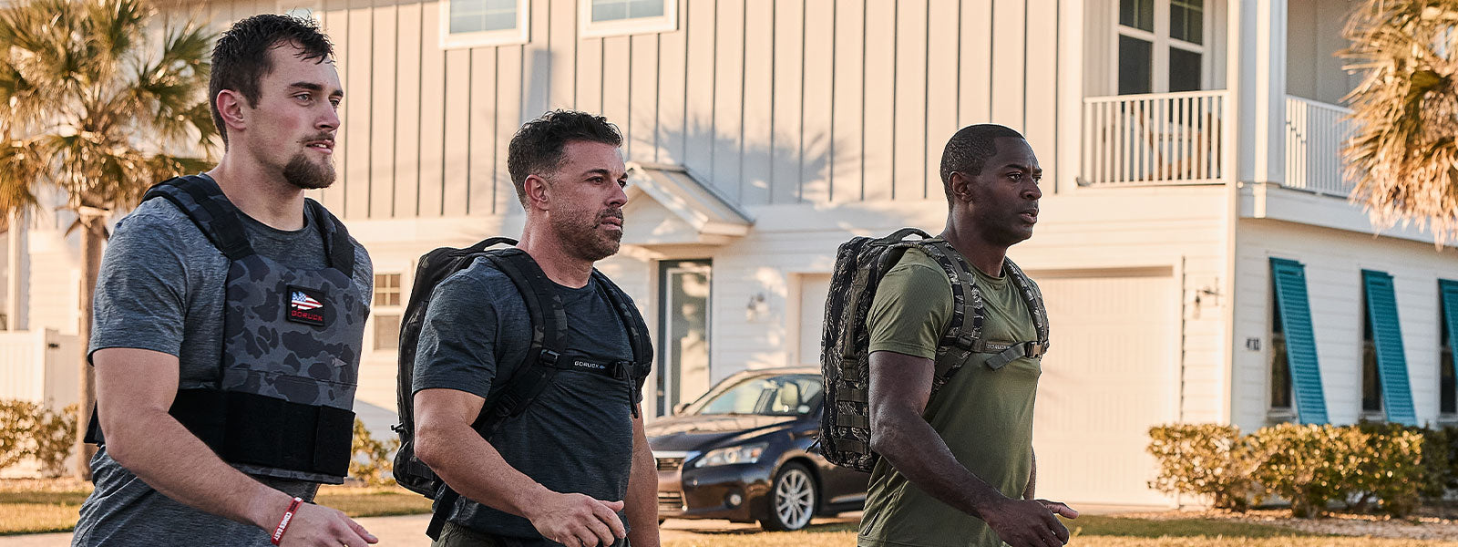 Three men with weighted vests walk outdoors near houses on a sunny day.