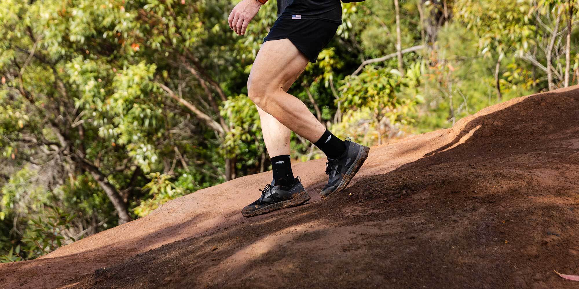 A person in athletic shorts and shoes hikes up a dirt trail surrounded by greenery.