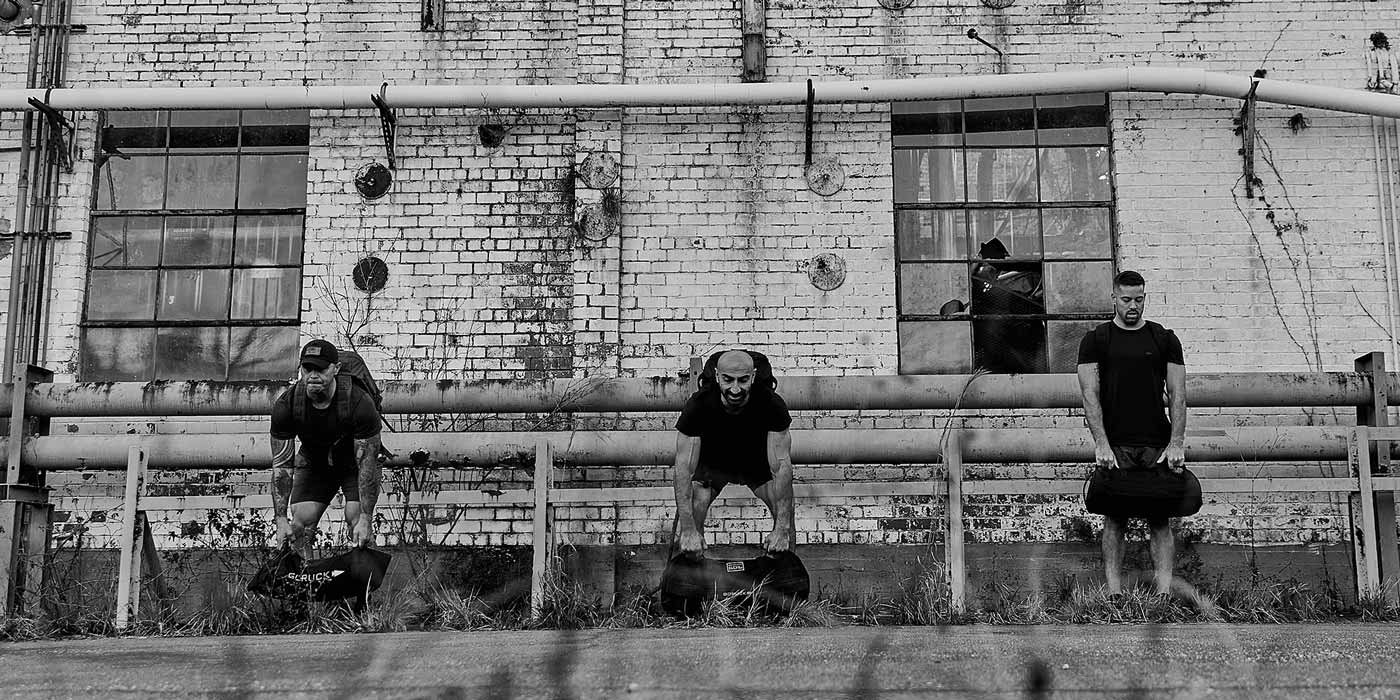 Three people lifting sandbags in front of a weathered brick wall with large windows in a black-and-white photo.