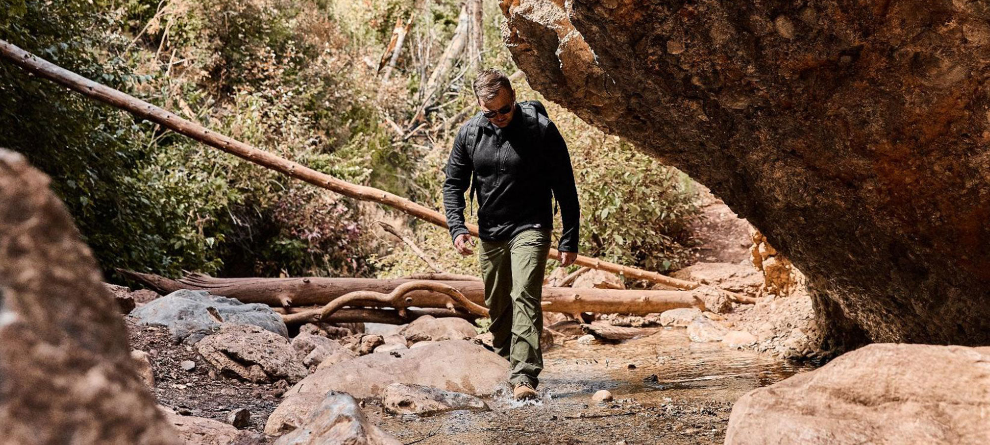 A person walks over a rocky stream under a large boulder in a forest setting.