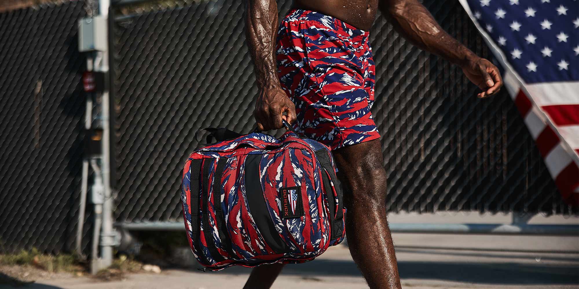 Person in red, white, and blue shorts walks with a matching bag, American flag visible in the background.