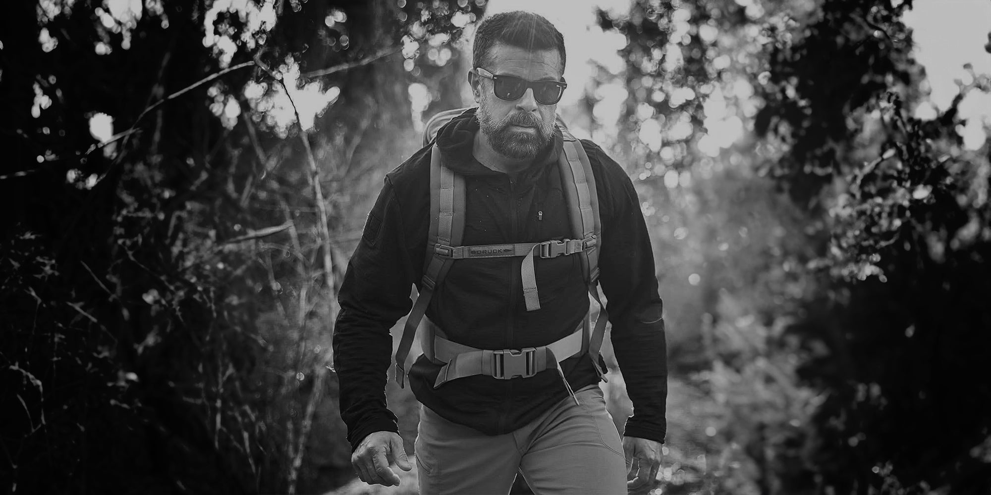 Man hiking through rocky terrain in a forested area, wearing a backpack. Black and white image.