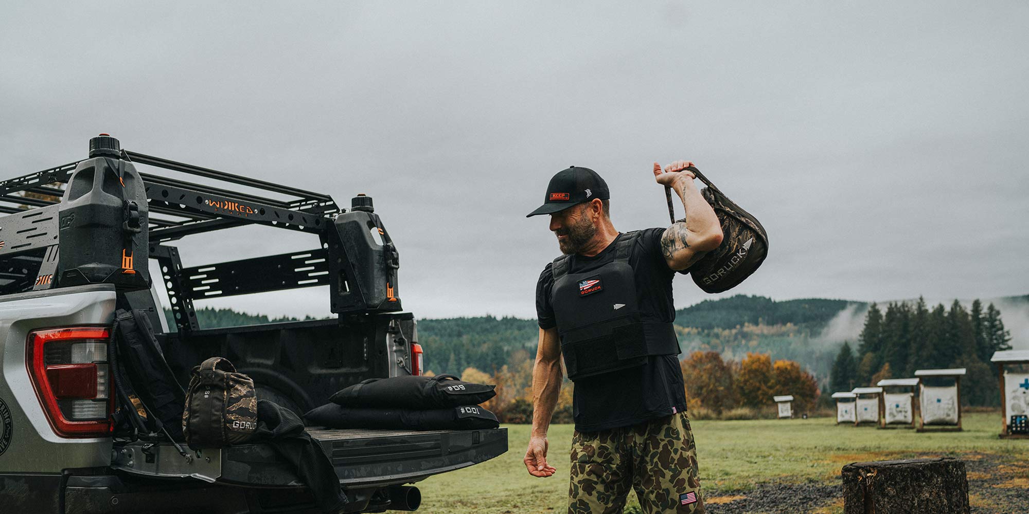 Man in tactical gear lifts a sandbag near a truck on a grassy field, with forested hills in the background.