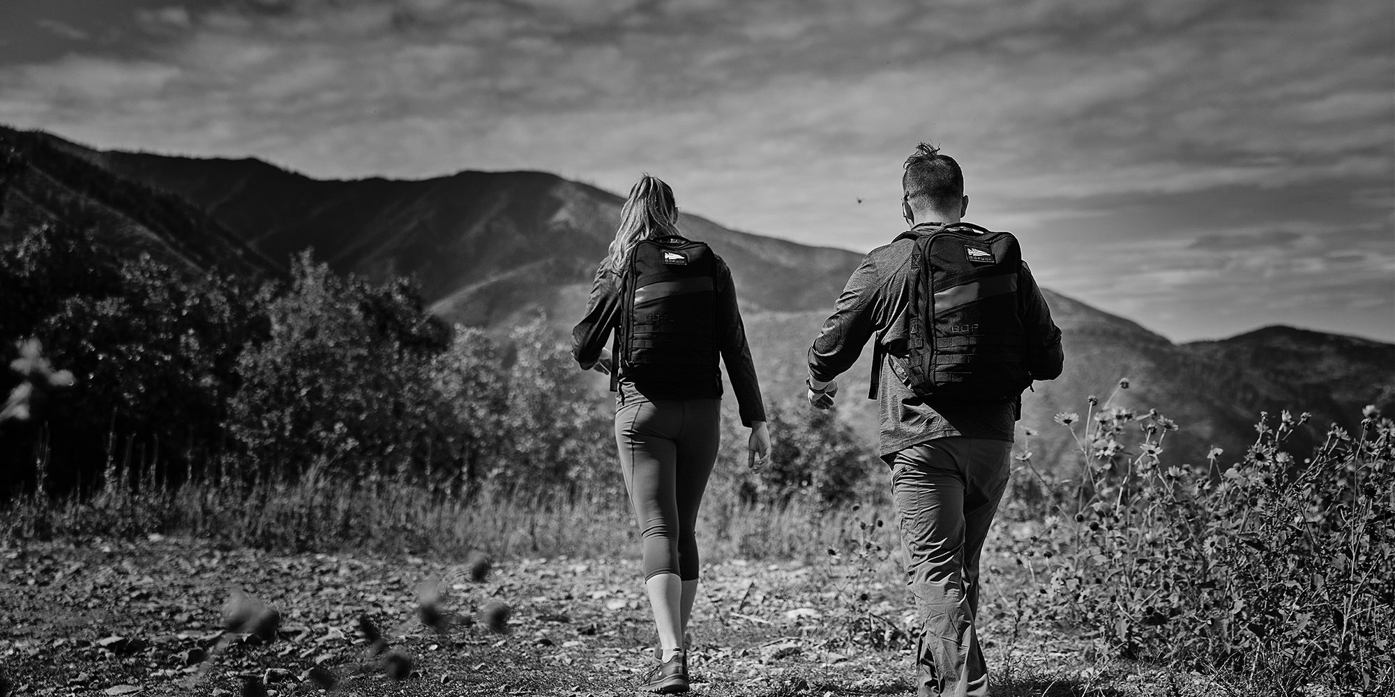 Two men with backpacks walking and running outdoors, wearing camouflage shorts and dark T-shirts.