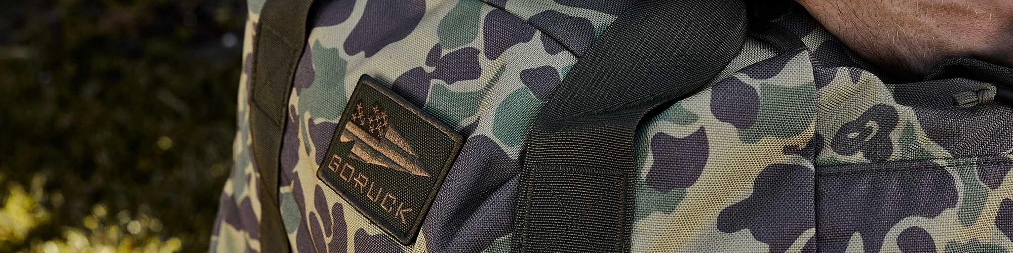 Close-up of a green camouflage backpack with a patch featuring an American flag and the text "GORUCK".