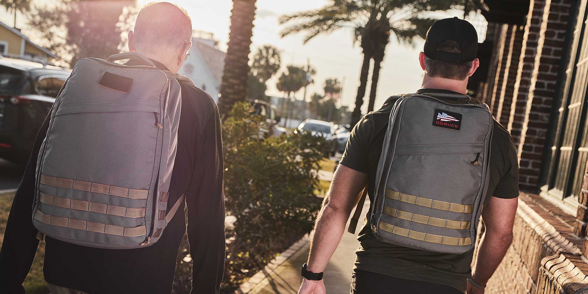 Two people wearing backpacks walk down a sunny street lined with palm trees, with cars parked nearby.