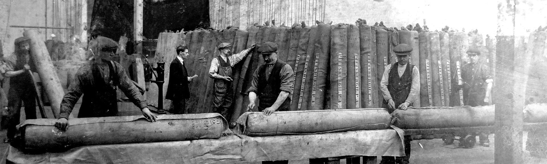 Black and white photo of workers in a factory rolling large fabric pieces on wooden tables.