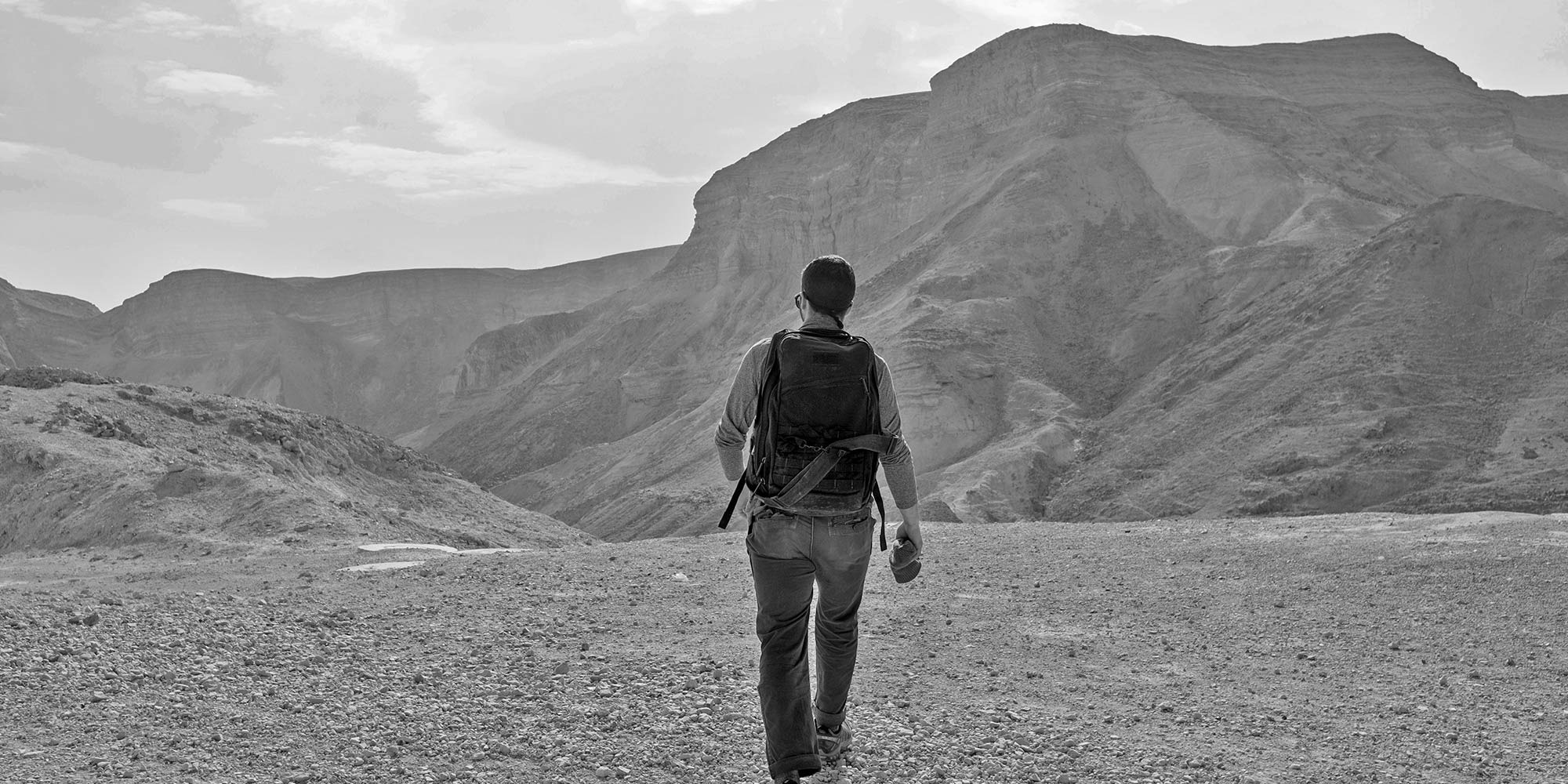 A person with a backpack walks toward rocky mountains under a cloudy sky.