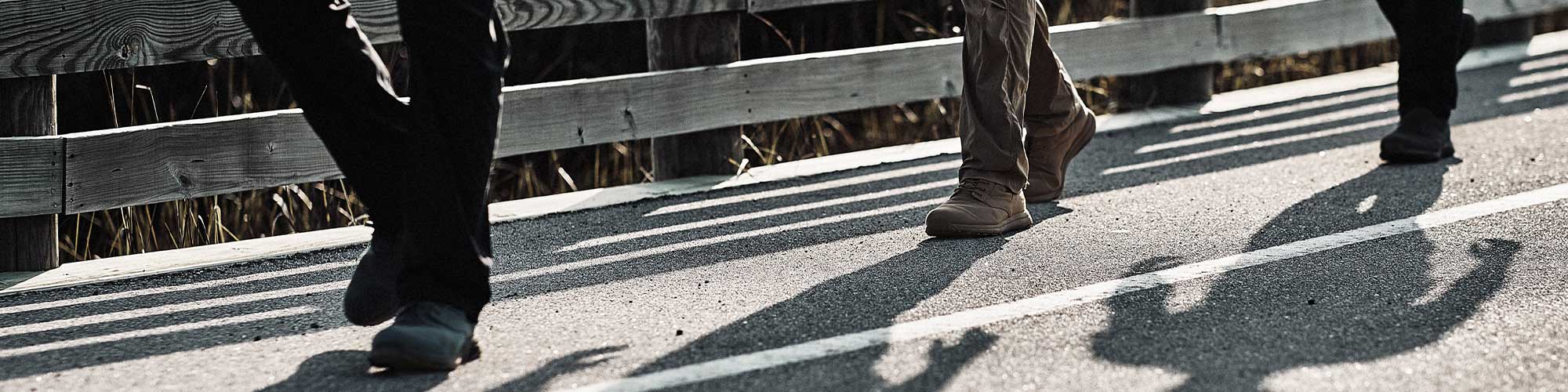 Two people walking on a paved path beside a wooden fence, casting long shadows on the ground.