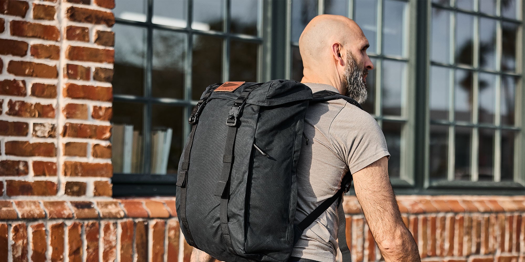 Bald man with beard wearing a gray t-shirt and black backpack, standing in front of a brick building with windows.