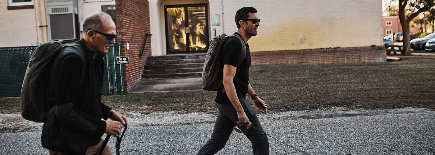 Two men wearing backpacks and sunglasses walk dogs on leashes past a building.