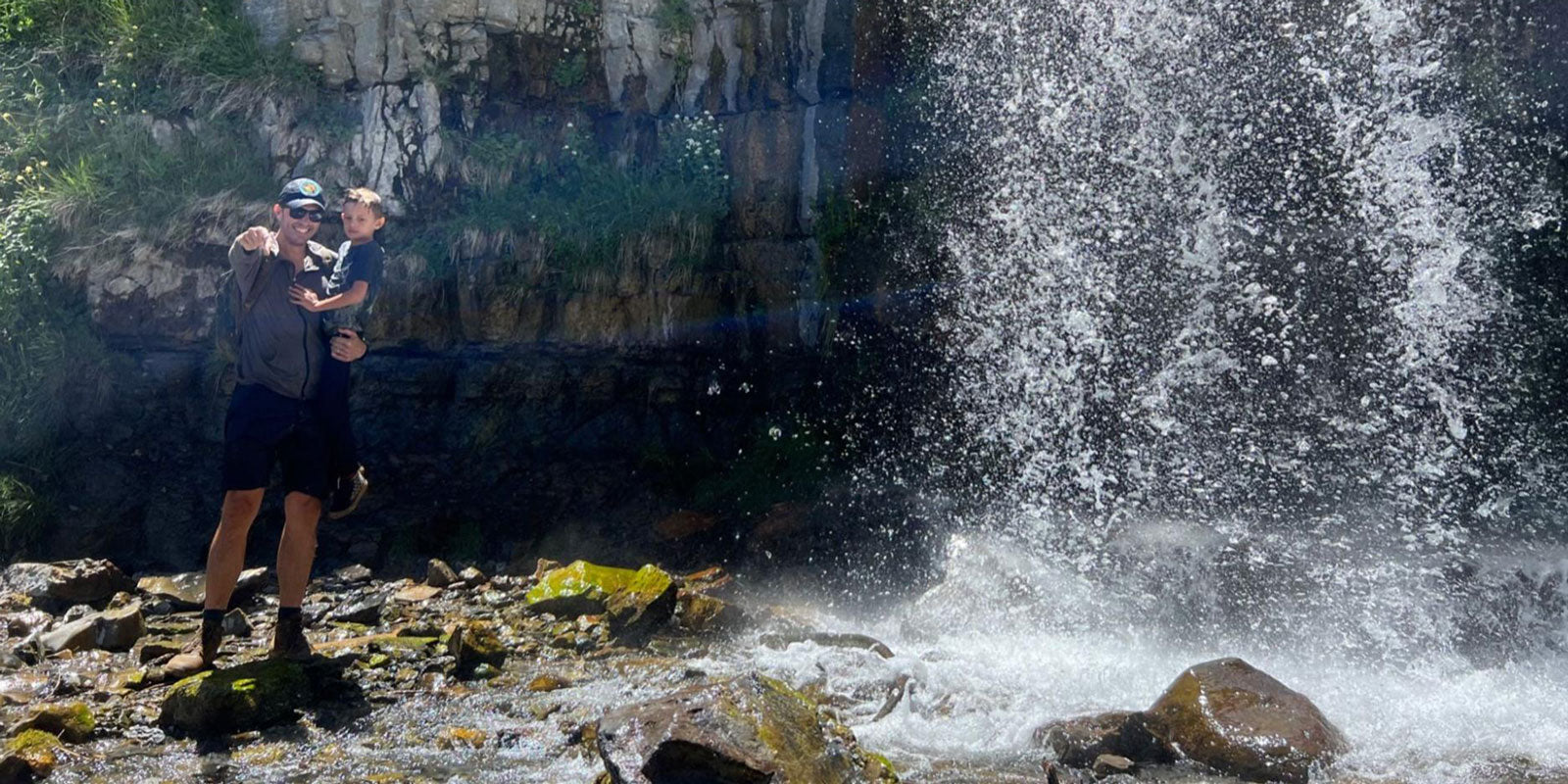 Two people stand near a cascading waterfall, surrounded by rocks, in a lush outdoor setting.