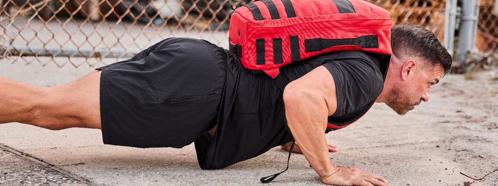 Man doing push-ups on concrete, wearing a red weighted backpack and black athletic clothes. Chain-link fence in background.