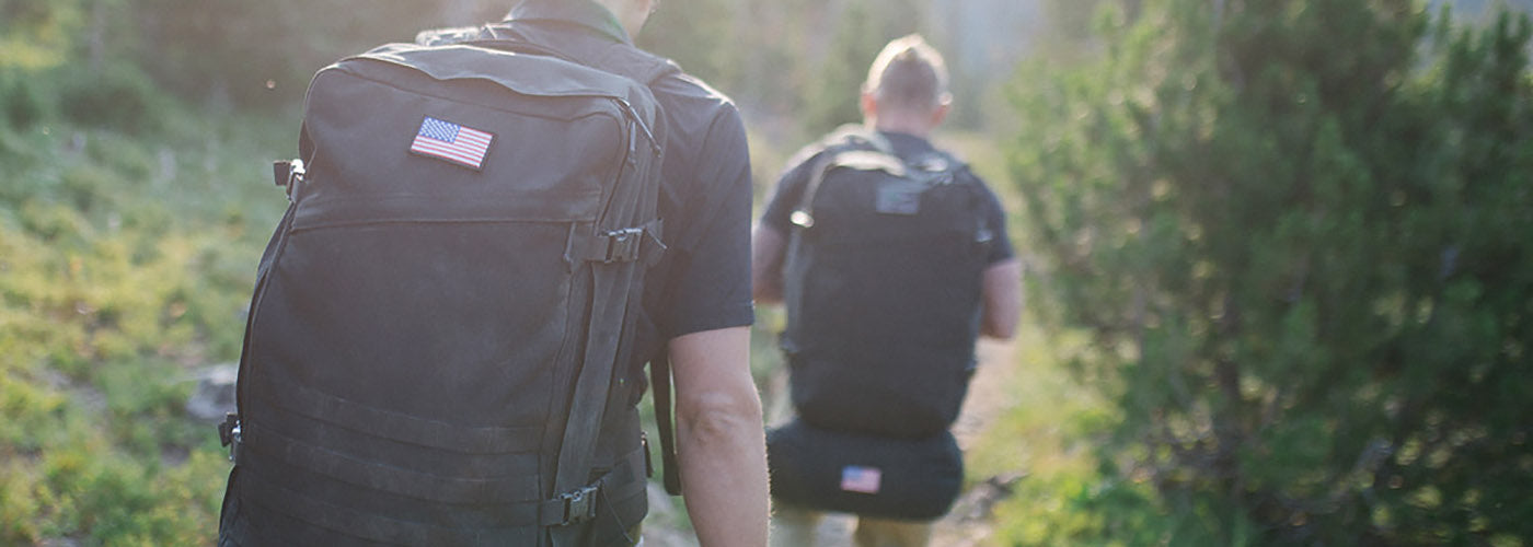 Two people hiking on a trail with backpacks featuring American flags in a sunlit forest.