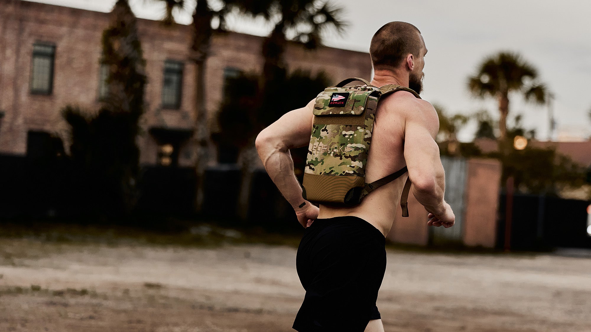 Man jogging outdoors with a camouflage backpack, wearing black shorts. Buildings and palm trees in the background.