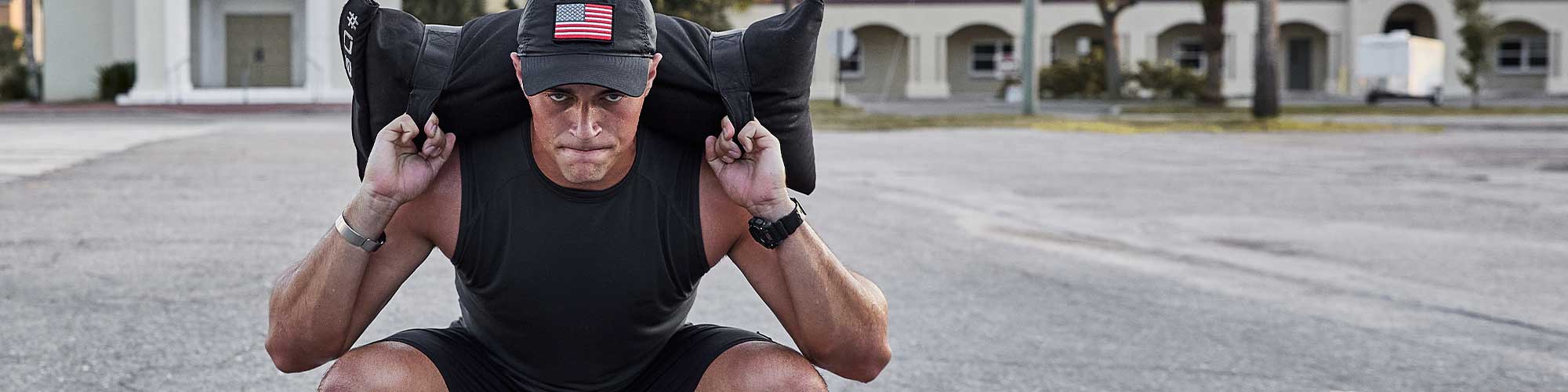 A man squats outdoors, holding a sandbag on his shoulders, wearing a cap with a USA flag patch.