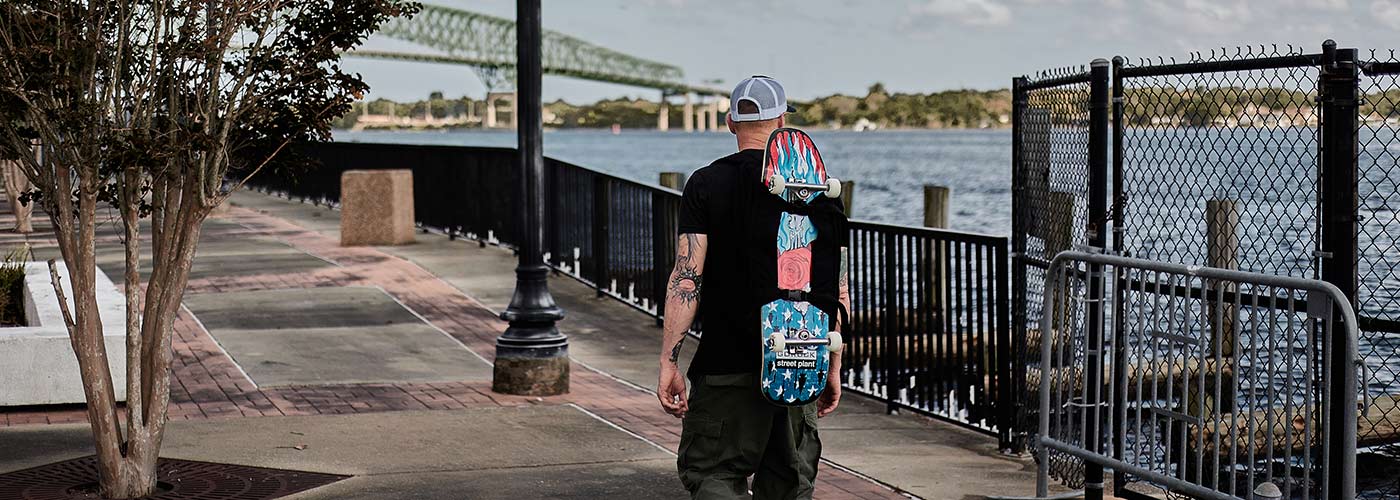 Person walking along a waterfront path, carrying a skateboard, with a bridge in the background.