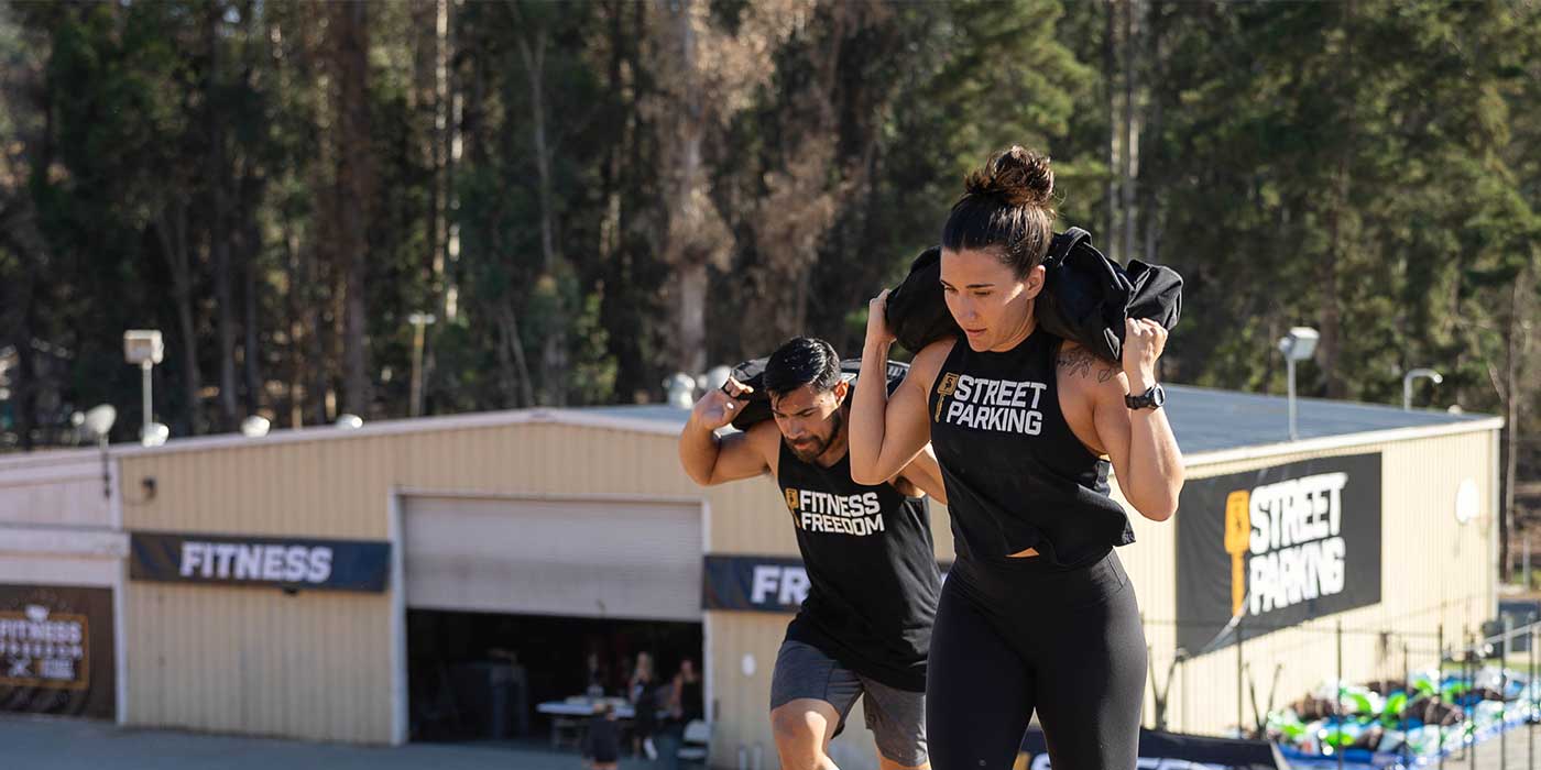 Two people exercise outside, carrying sandbags on their shoulders near a fitness facility.