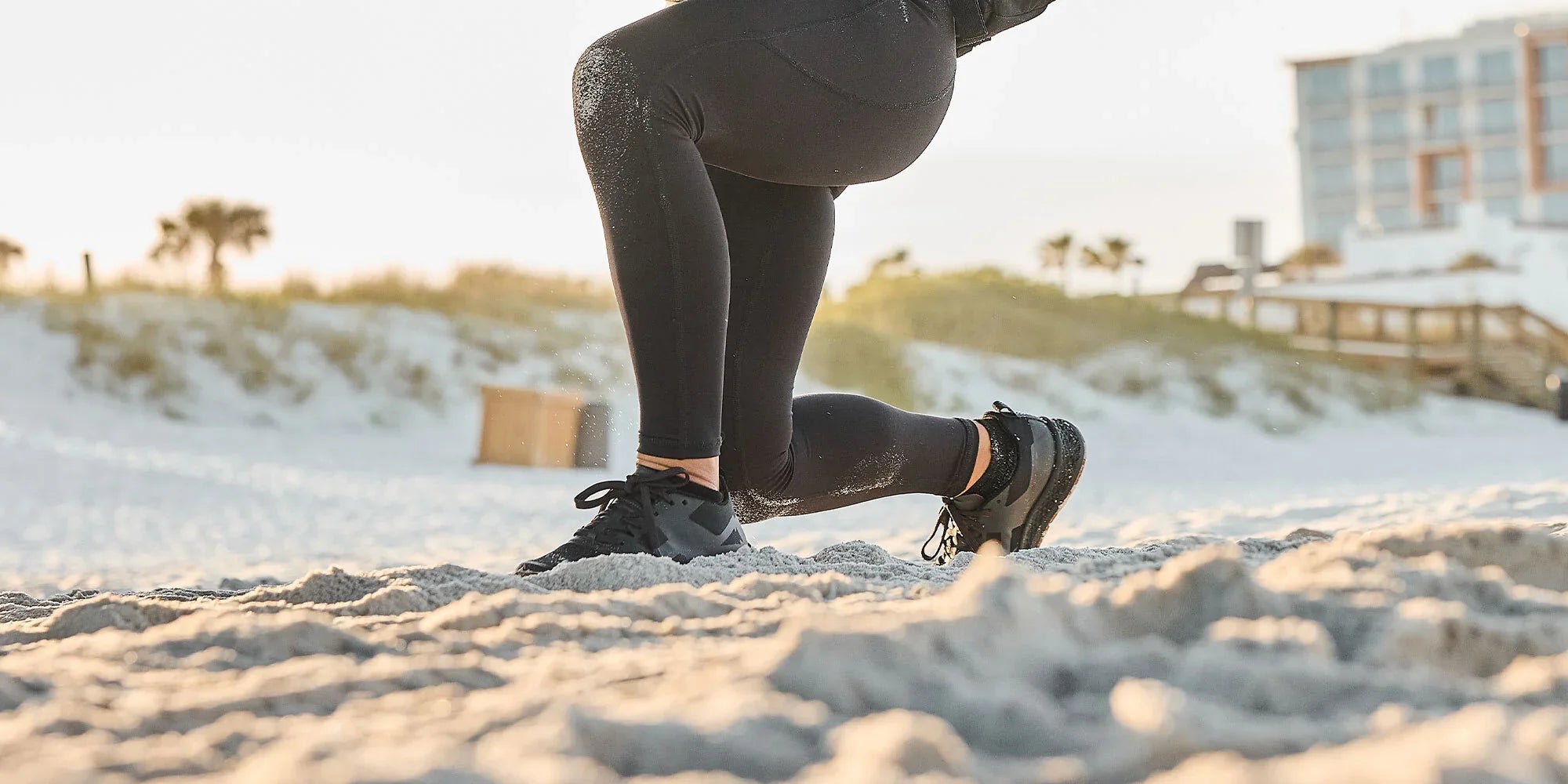 Person in black leggings doing lunges on a sandy beach with buildings and plants in the background.