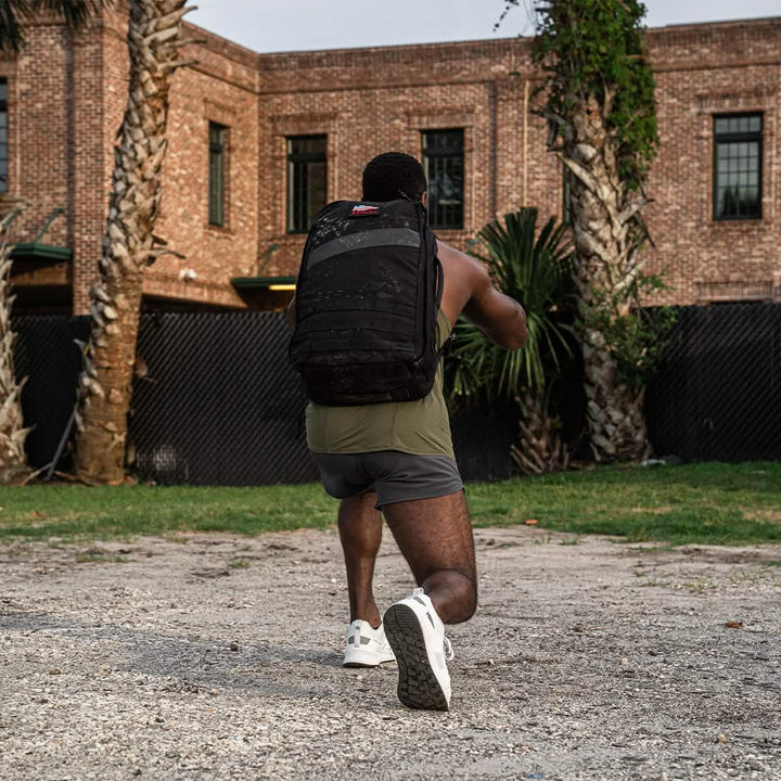 A man in workout gear lunges outdoors with his GORUCK Rucker 4.0 backpack, set against a backdrop of a brick building and swaying palm trees.