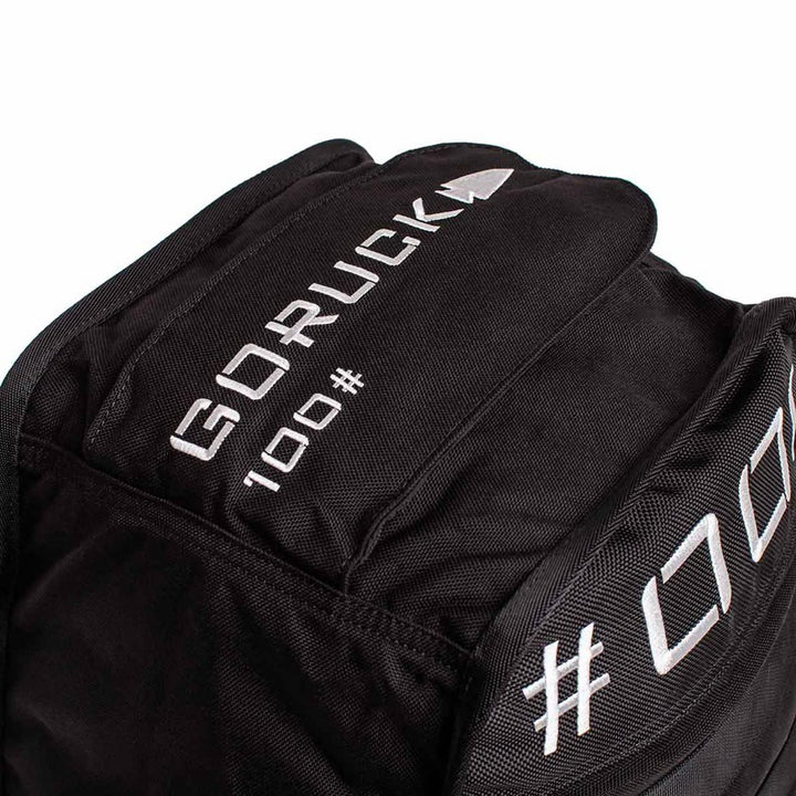 Close-up of a black backpack with white text reading "GORUCK." The backpack features a multilayer closure system and durable fabric, suggesting it's designed for heavy-duty use reminiscent of carrying GORUCK's Sand Tombstones.