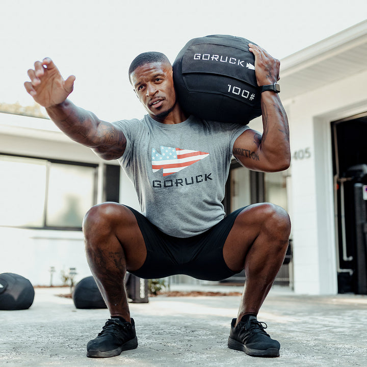 A man squats with a 100 lb Sand Medicine Ball from GORUCK on his shoulder, showcasing core stability outside a building, as if bringing the intensity of a home gym workout to the outdoors.