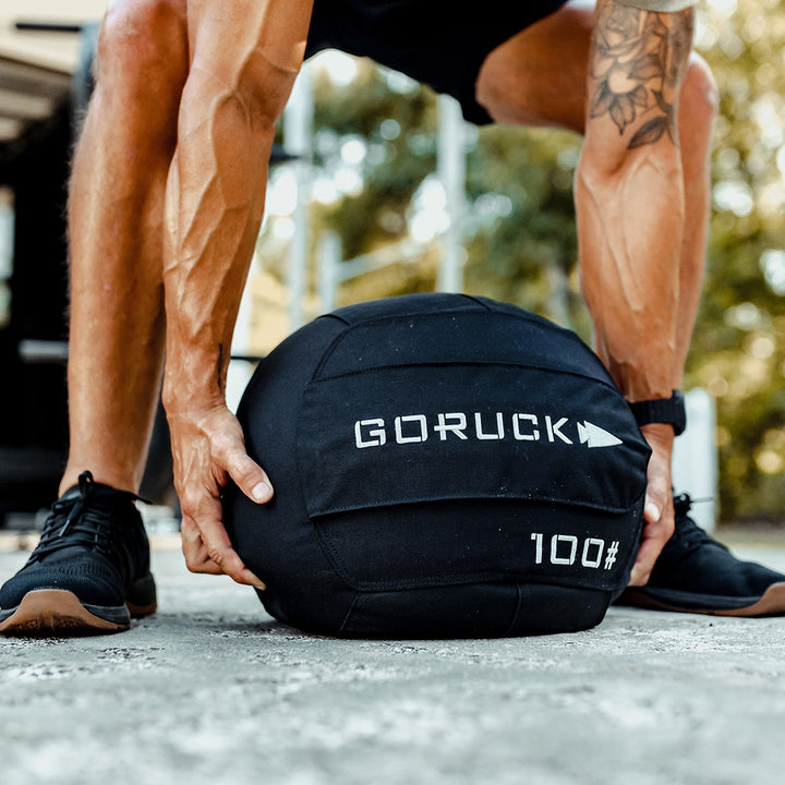 Outdoors, a person grips a GORUCK Sand Medicine Ball in black, enhancing core stability like a versatile 100-pound sandbag from their home gym.