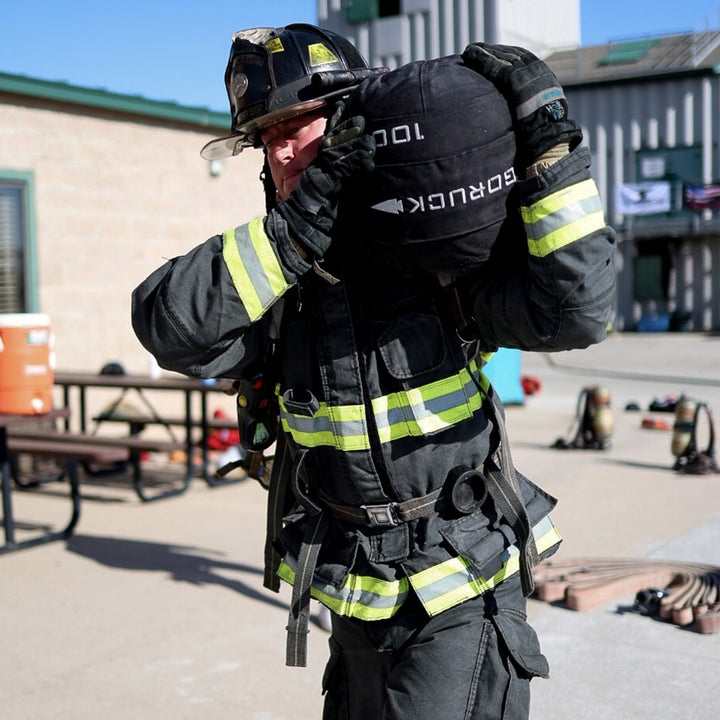 A firefighter in full gear carries a heavy black equipment bag outdoors near the training area, where GORUCK Sand Medicine Balls and other gear are used to enhance core stability. Equipment is visible in the background.