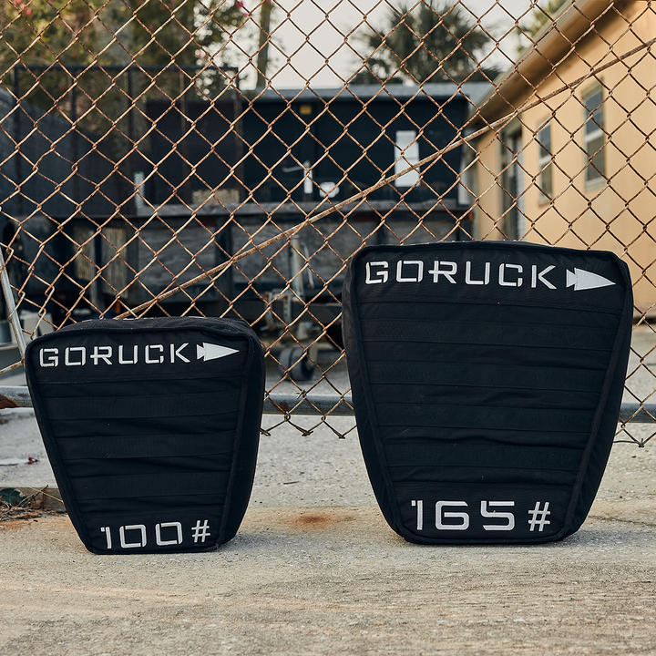 Two black Sand Tombstones from GORUCK, reminiscent of a Husafell Sandbag, rest against a chain-link fence. They are marked with arrows and weights of 100 and 165. In the background, an industrial landscape dotted with buildings and trees provides the perfect setting for challenging loaded carries.