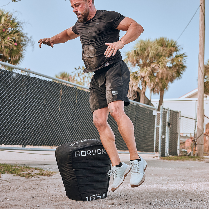 A man exercises outdoors in a black t-shirt and shorts, jumping onto a black Sand Tombstones by GORUCK, which features a multilayer closure system. Palm trees and a chain-link fence form the background, enhancing the rugged setting perfect for loaded carries or using the Husafell Sandbag.