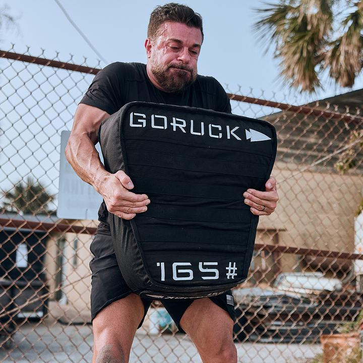 A man is outdoors, struggling to lift a large black padded Sand Tombstone from GORUCK, weighing 165 lbs, reminiscent of a modern Husafell Sandbag challenge. He is wearing a dark shirt, with a chain-link fence and trees and buildings visible in the background.