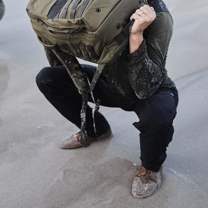 On a sandy beach, a person squats, lifting a military-style backpack overhead, their GORUCK MACV-1 Hi-Speed Mid Top boots firmly planted in the sand.