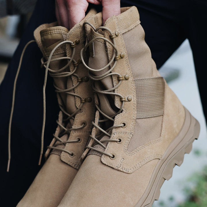 A close-up of a pair of GORUCK MACV-2 High Top boots in coyote color, showcasing laces and rugged soles. The boots feature a triple compound outsole and are being held by a person's hand against a blurred background.