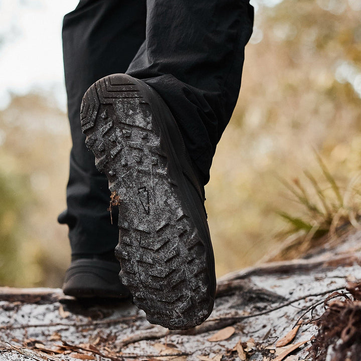 A close-up of the MACV-2 - Mid Top - Black boot from GORUCK stepping on a muddy, uneven trail. The triple compound outsole is detailed with dirt and grass. The background is blurred, revealing subtle hints of green foliage and brown earth.