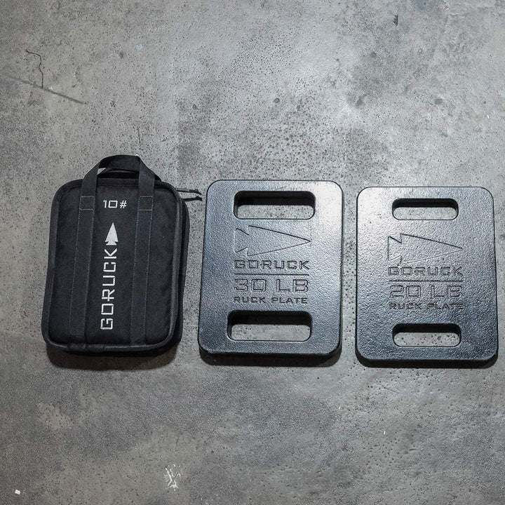 Two portable Sand Ruck Plates and a GORUCK weight bag rest on a concrete floor, ready for your next rucking adventure.