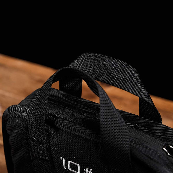 Close-up of a black fabric bag with handles, resting on a wooden surface against a black background, perfect for carrying your portable GORUCK Sand Ruck Plates for rucking adventures.