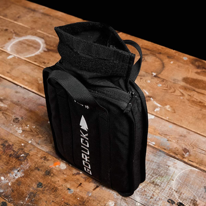 A black GORUCK Sand Ruck Plate bag rests on a wooden table, partially open with zippers visible, ready for your next rucking adventure.