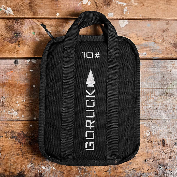 A black GORUCK Sand Ruck Plate bag with handles rests on a wooden surface, prominently displaying its white logo and the number "10" with an arrow design. This bag is perfectly designed to hold a portable Ruck Plate for your rucking adventures.