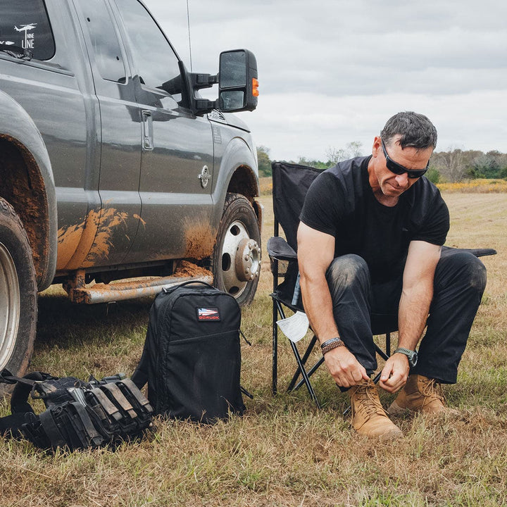 A man sits by a muddy truck wearing sunglasses, tying his boots. Beside him lies a GORUCK Bullet Ruck Double Compartment made of ripstop nylon, full of gear.